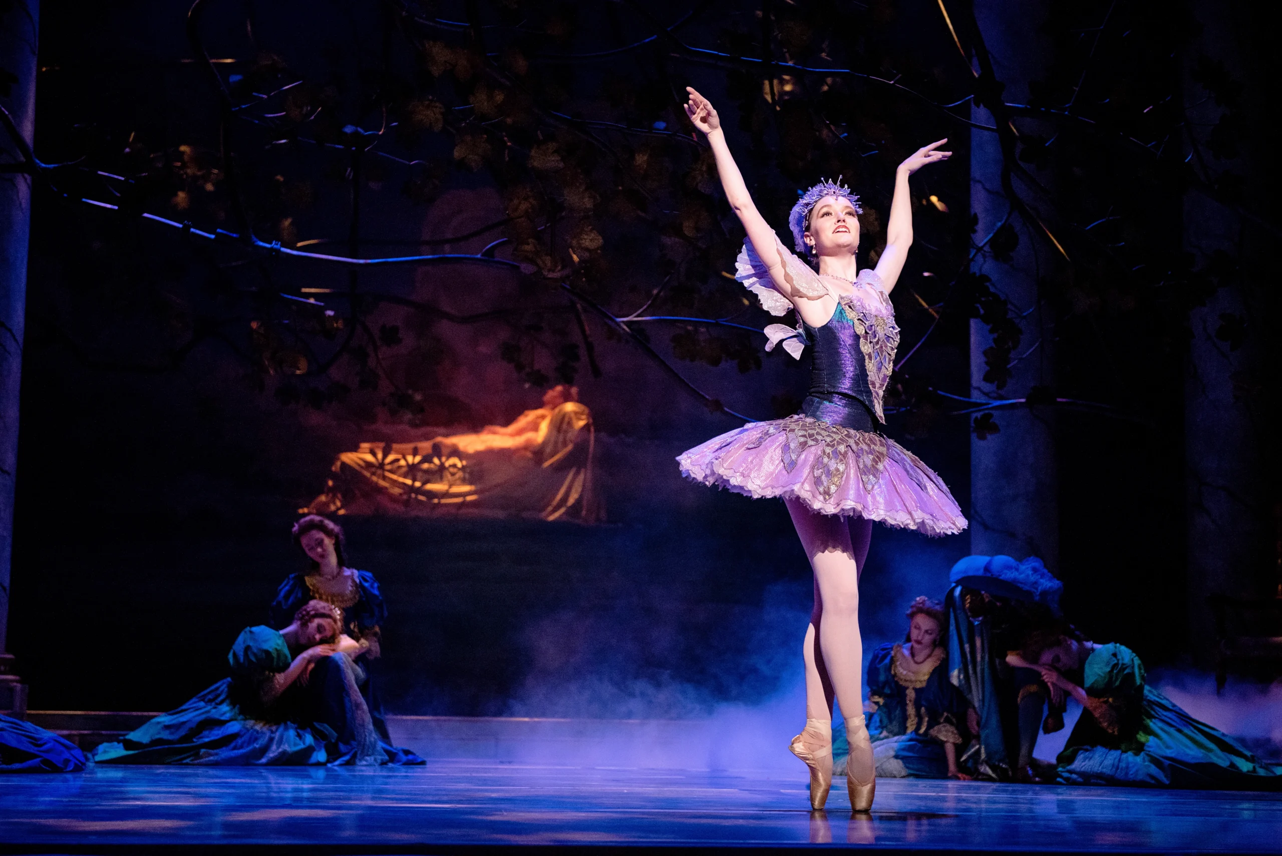 Sarah Lapointe performs onstage in a purple tutu and crown. She smiles and stands in sus-sous on pointe with her right foot front and her arms lifted up into a V shape. Behind her the stage is dark and lit in purple, with a warm spotlight on a dancer as Sleeping Beauty lying down on a bed in the background. Two clusters of dancers sit on the floor as if asleep.