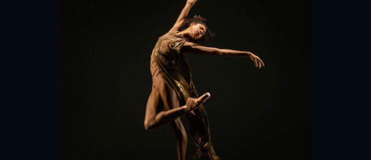Adji Cissoko poses in a dramatic contemporary retire against a pitch black background. She wears a flowing gold dress and flesh-tone pointe shoes, leaning back and raising her arms in abandonment.