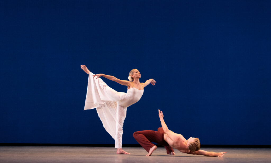 Dawn Atkins stands in a high arabesque onstage, on flat. She wears a long, flowing white dress and a flower in her hair, which is pulled into a low bun. A male dancer poses in an extreme hinge position on his back on the floor in front of her.