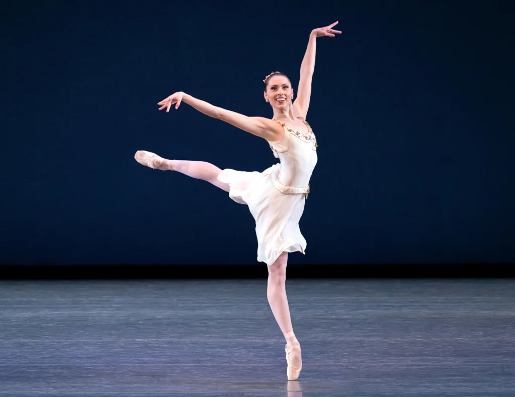 Isabella LaFreniere, wearing a huge smile, does a piqué in third arabesque with her left leg raised. She looks out to the audience, holding her left arm high, and wears a knee-length, white dance dress, pink tights, and pointe shoes.