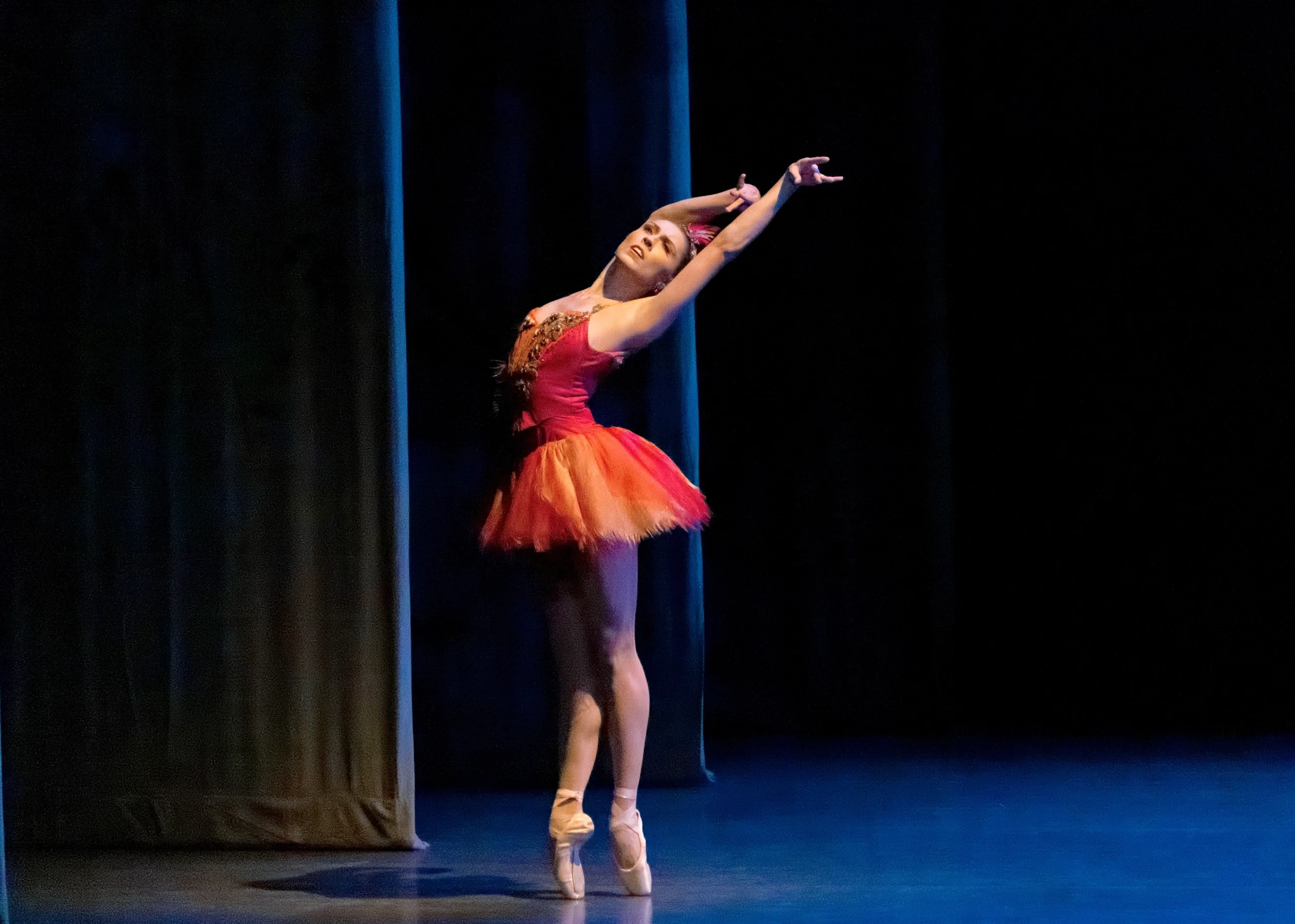 Wearing a short red and gold tutu, Isabella LeFreniere poses in sus-sous effacé on pointe during a performance of Firebird. She stands close to the wings on stage right and lifts her arms high over her head, sweeping them to the left and arching her upper back.