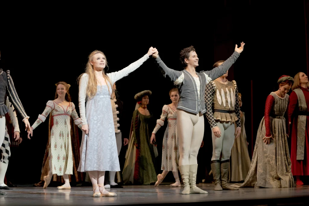 Sara Mearns and Guillaume Côté stand center stage and lift their arms to take a bow, joining hands. The supporting cast of dancers stand in lines behind them. Mearns wears a light gray empire-waisted dress, pink tights and pointe shoes; Côté wears a dark gray tunic, beige tights and beige boots, and a large smile on his face.
