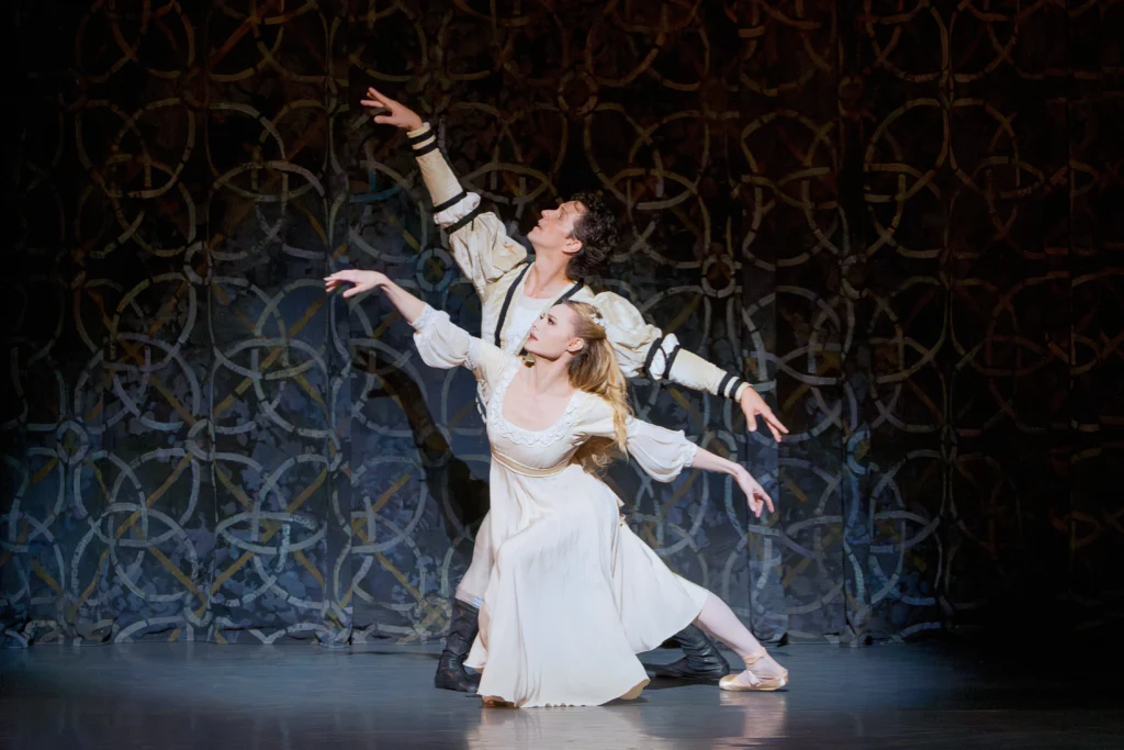 Guillaume Côté and Sara Mearns perform a scene from Romeo and Juliet in front of a patterned backdrop. They both take low lunges in fourth position facing stage right, their arms stretched out in a first arabesque line. Côté, standing directly behind Mearns and in a slightly higher lunge, wears an off-white tunic with puffed upper sleeves and dark trim. Mearns wears a cream-colored empire waist dress, pink tights and pointe shoes.