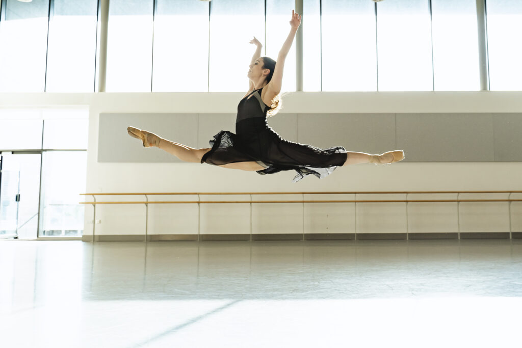 Alessandra Bramante flies across a brightly-lit dance studio in a saut de chat, her arms lifted in a high "V". She wears a long flowing black dress and ballet flats.