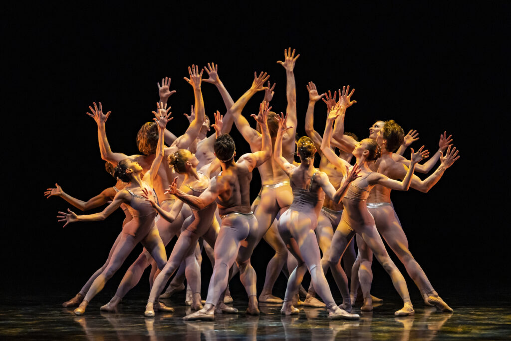 a large group of male and female dancers in grayish-nude unitards cluster in a group, each pointing their hips into the center of the cluster, in a lunge. they raise their arms, bent gently, to the sides and spread their fingers and palms wide.