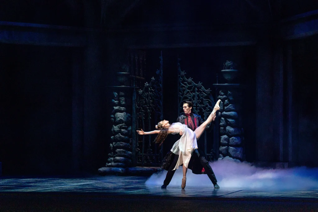 Cecliy Khuner and Barndon Ramey perform a scene from the ballet Dracula onstage. They dance in front of a old gothic gate with fog rolling in from stage left. Ramey, dressed as Dracula, holds Khuner by the waist as she stands on pointe and lays back into his arms, raising her left leg up in a high dévelloppé devant. She wears a white tattered dress and pointe shoes, and her hair long.