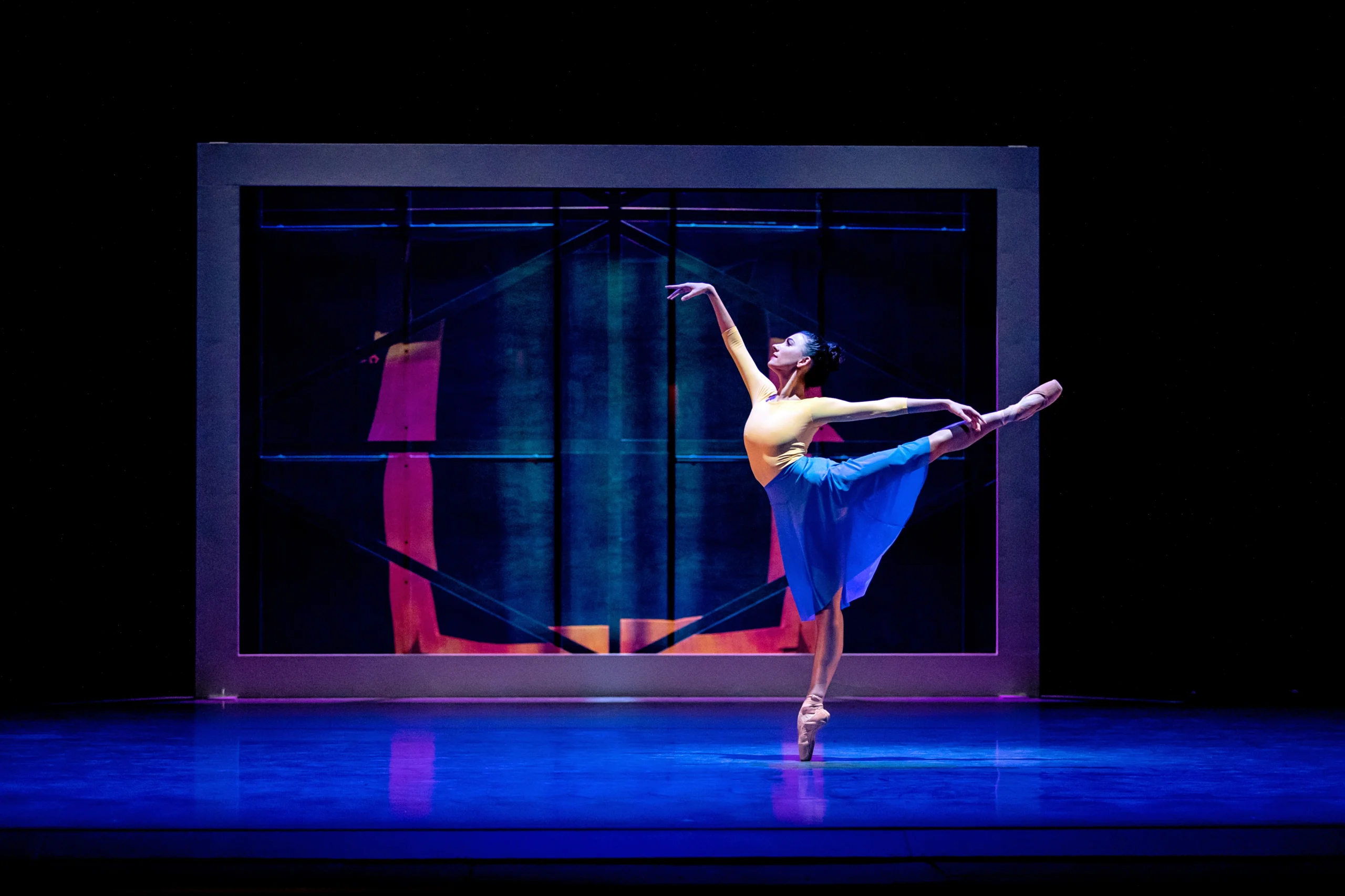 During a performance, Jasmine Jimison performs a piqué in first arabesque on pointe towards stage right. She wears a yellow long-sleeved top and bright blue skirt, and dances in front of a large, framed multicolored image.