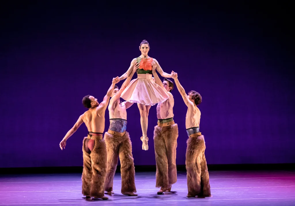 Four bare-chested men in brown, wooly chaps surround a ballerina and lift her up during a performance. The woman, who closes her legs in sus-sous with her feet pointed, wears pointe shoes and a knee-length dress with a green and coral bodice and pink, poofy skirt. She smiles directly at the audience as two men hold her hands and the other two lift her from the armpits. They perform in front of a purple backdrop.