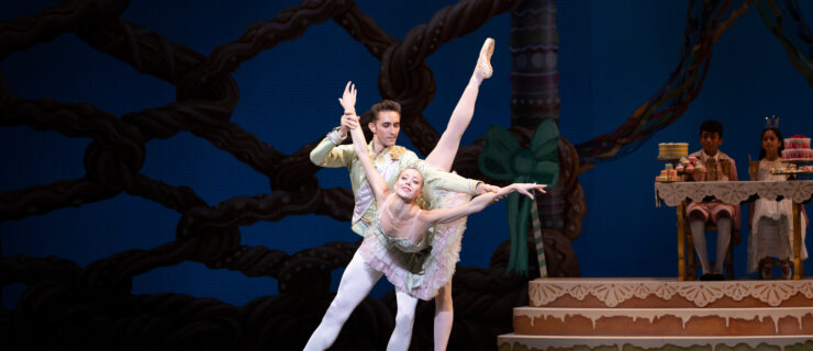 Dawn Atkins takes a deep penché arabesque in croisé on pointe with her right leg raised and her arms out to the side. Stanislav Olshanskyi stands behind her and slightly to her left in a tendu second, holding onto her wrists to help support her. Atkins wears a mint green tutu with a pink skirt, while Olshanskyi wears white tights and a mint green tunic. Behind them onstage, a boy and girl in costumes sit at a table piled with sweets and cakes, watching.