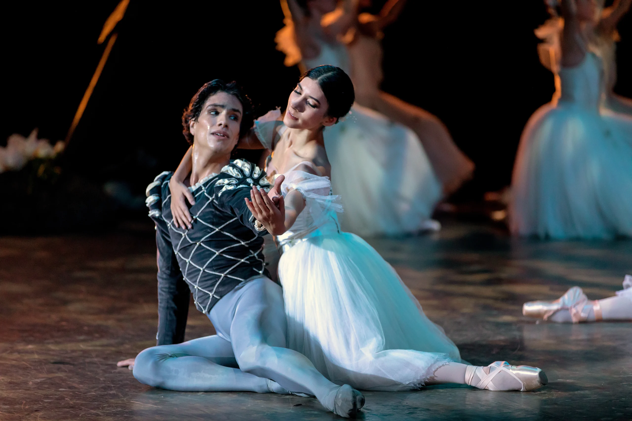 During a performance of the ballet Giselle, Esteban Hernández sits on the ground, legs extended to his left, and props himself up on his right hand. He looks desperately back at Jimison, who sits behind him, holding him around his right shoulder and under his extended left hand. Jimison wears a long, white Romantic tutu, tights and pointe shoes, while Hernandez wears gray tights and a dark gray, velvet tunic.
