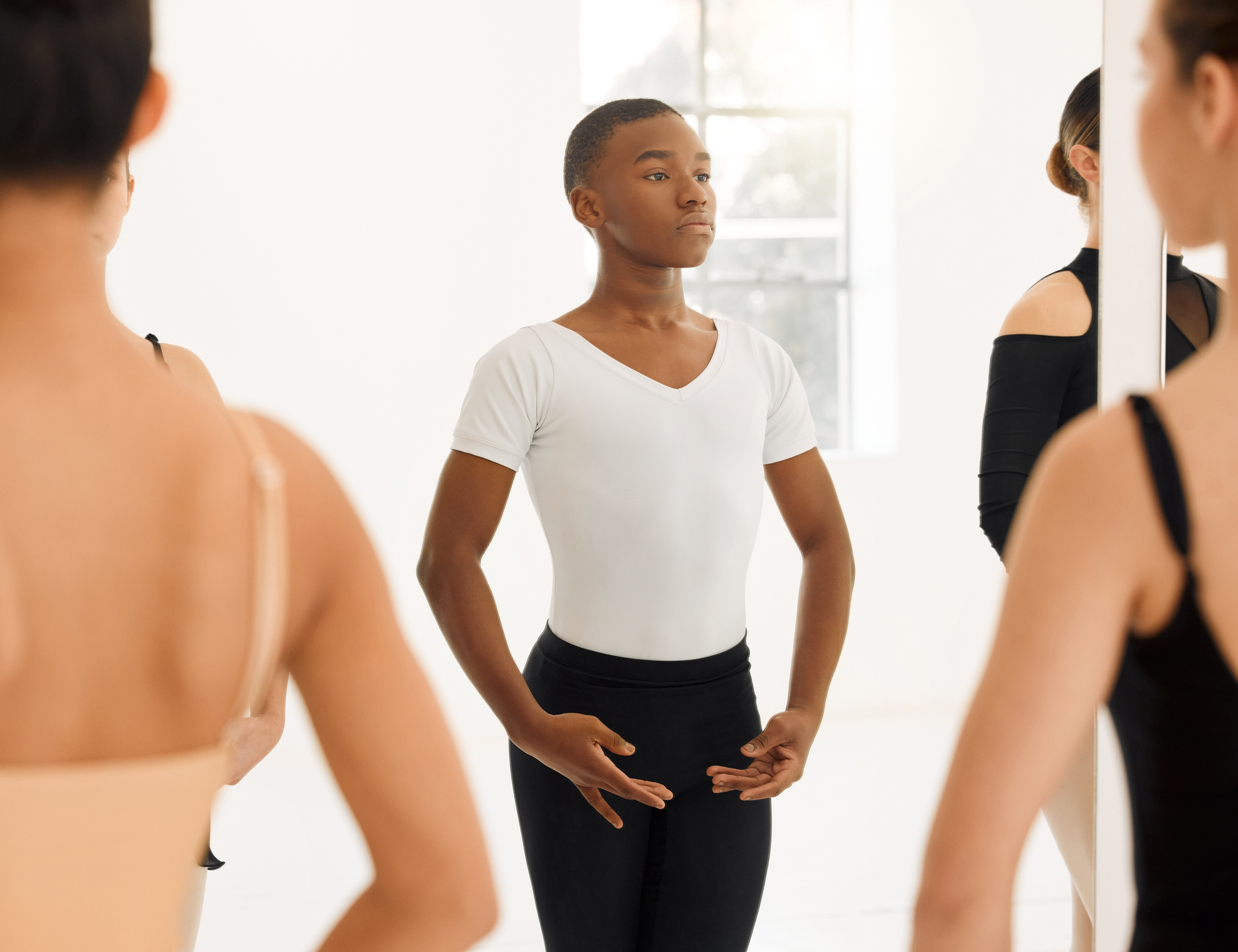 A teenage male ballet student is shown from the knees up in a large, brightly lit dance studio. He stands in fifth position with his arms en bas, and wears a white V-neck T-shirt and black tights. He looks into the mirror with focus.