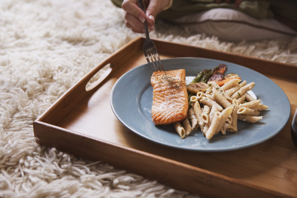A meal of salmon with pasta and vegetables is served on a blue plate atop a wooden tray. A hand holds a fork reaching toward the salmon, and a fuzzy white carpet is underneath everything.