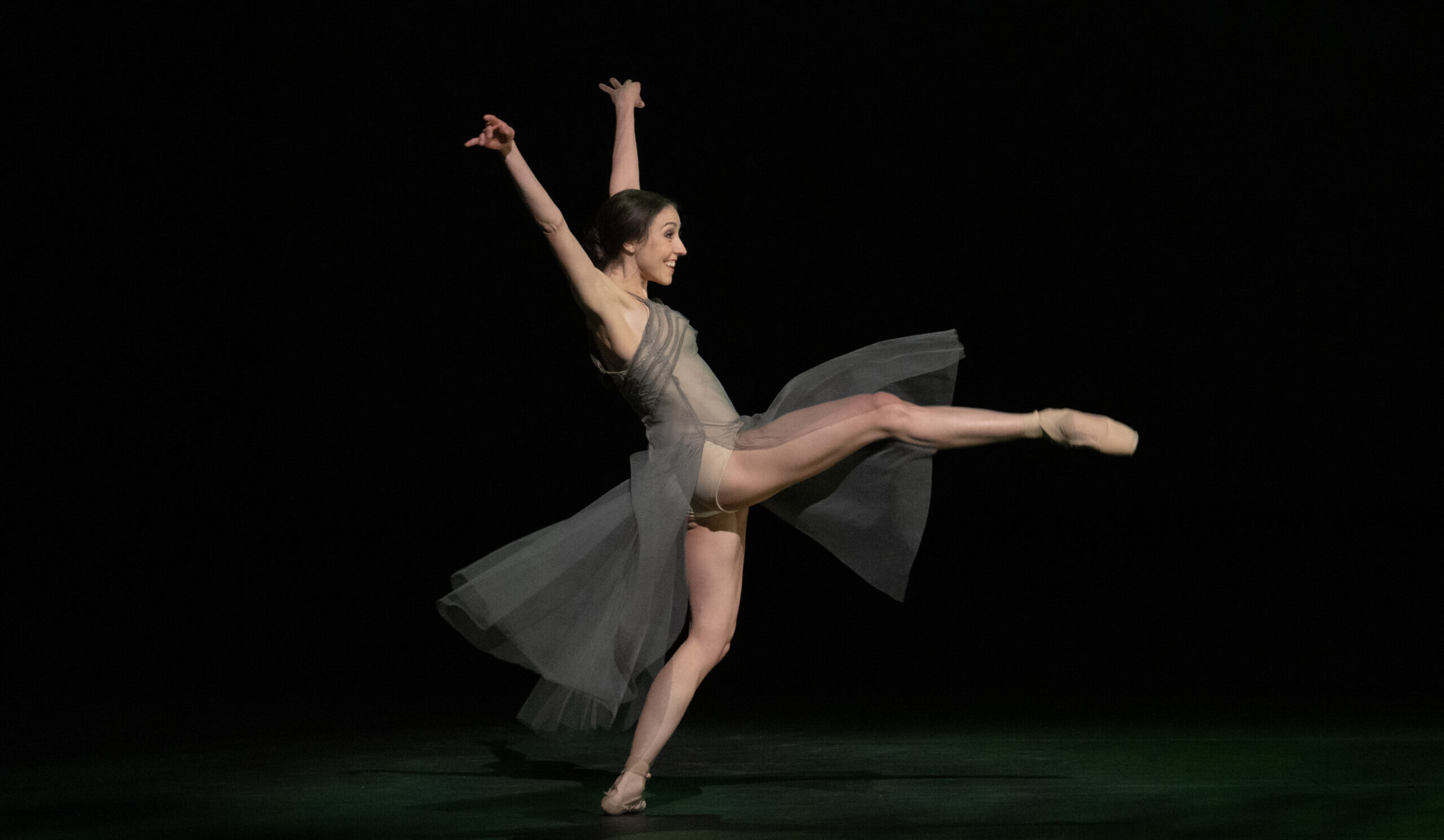 Ballet news. On a dark stage, Isabella Gasparini smiles as she does an attitude devant in a shallow lunge, arms lifted up in a "V" overhead. She wears a sheer taupe dress and pointe shoes.