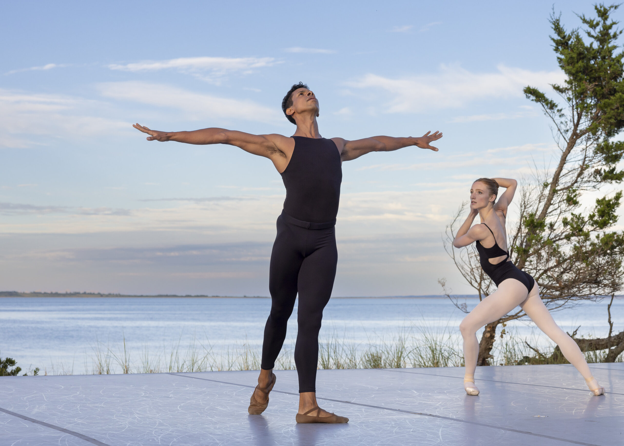 Jose Sebastian and Catherine Hurlin perform on a beautiful outdoor stage in front of a lake and blue sky at Hamptons Dance Project. Sebastian wears a black unitard, and Hurlin wears a black leotard with pink tights and pointe shoes.