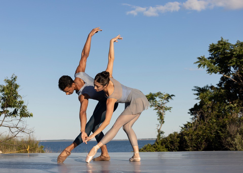 Jose Sebastian and Lauren Bonfiglio mirror each other as they perform outside at Hamptons Dance Project, in front of a lake and blue sky. They lunge in tendu devant, shown in profile, with their torsos twisting as they bend forward to hold hands by their feet and extend their back arms on the upward diagonal.