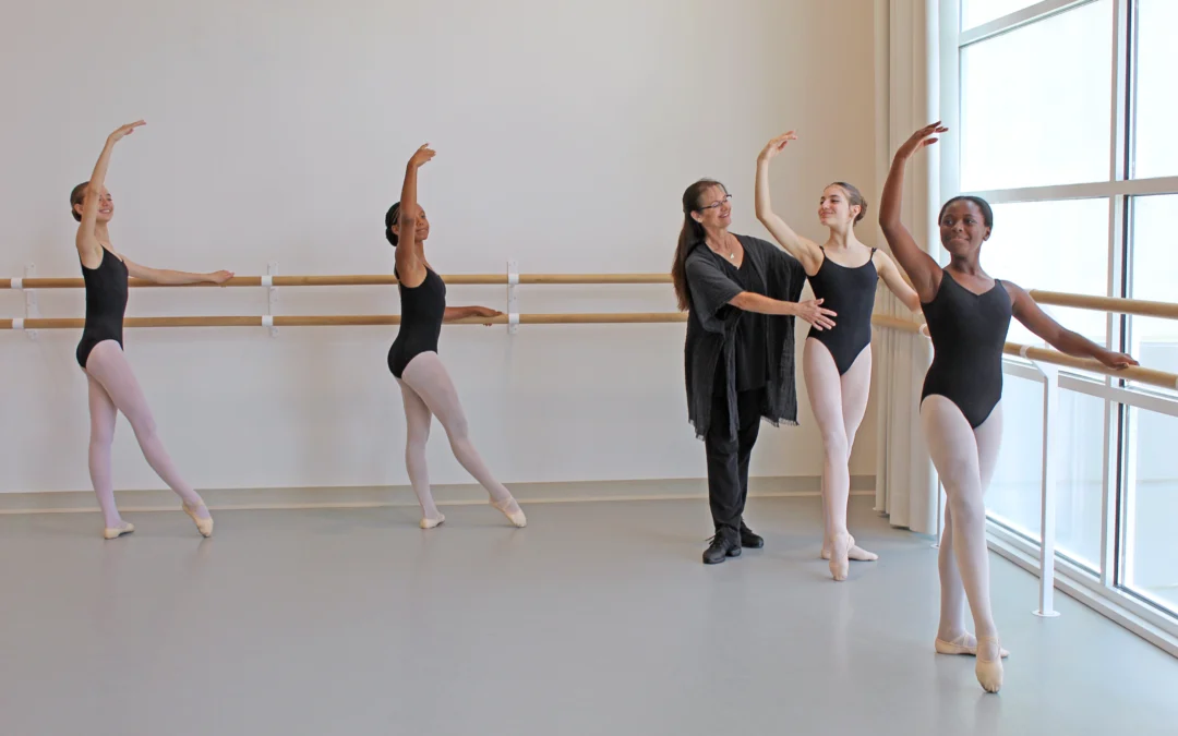 Kim Marsh, a ballet teacher, stands in the corner of a ballet studio and adjusts the hips of a female student practicing tendus at the barre. One student stands in front of her and two others on the opposite wall, doing the same exercise. Alll of the dancers wear black leotards, pink tights and pink ballet slippers. Marsh wears black pants and dance sneakers, a black V-neck shirt and a long, loose-fitting cardigan sweater.