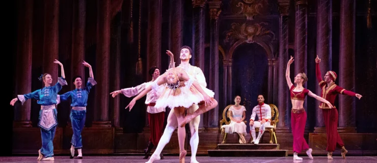 A large cast of dancers poses during a performance of Nutcracker. A ballerina portraying the Sugar Plum Fairy and her male partner stand center stage—he holds her waist as she stands facing him on pointe and does a deep backbend with her left leg in passé. She wears a pink tutu, brown tights and brown pointe shoes, while he wears white tights and slippers, and a white tunic. A male and female dancer sit in gilded chairs upstage center, while other dancers in various costumes frame the Sugar Plum Fairy and her cavalier in a V shape formation, posed in B plus.