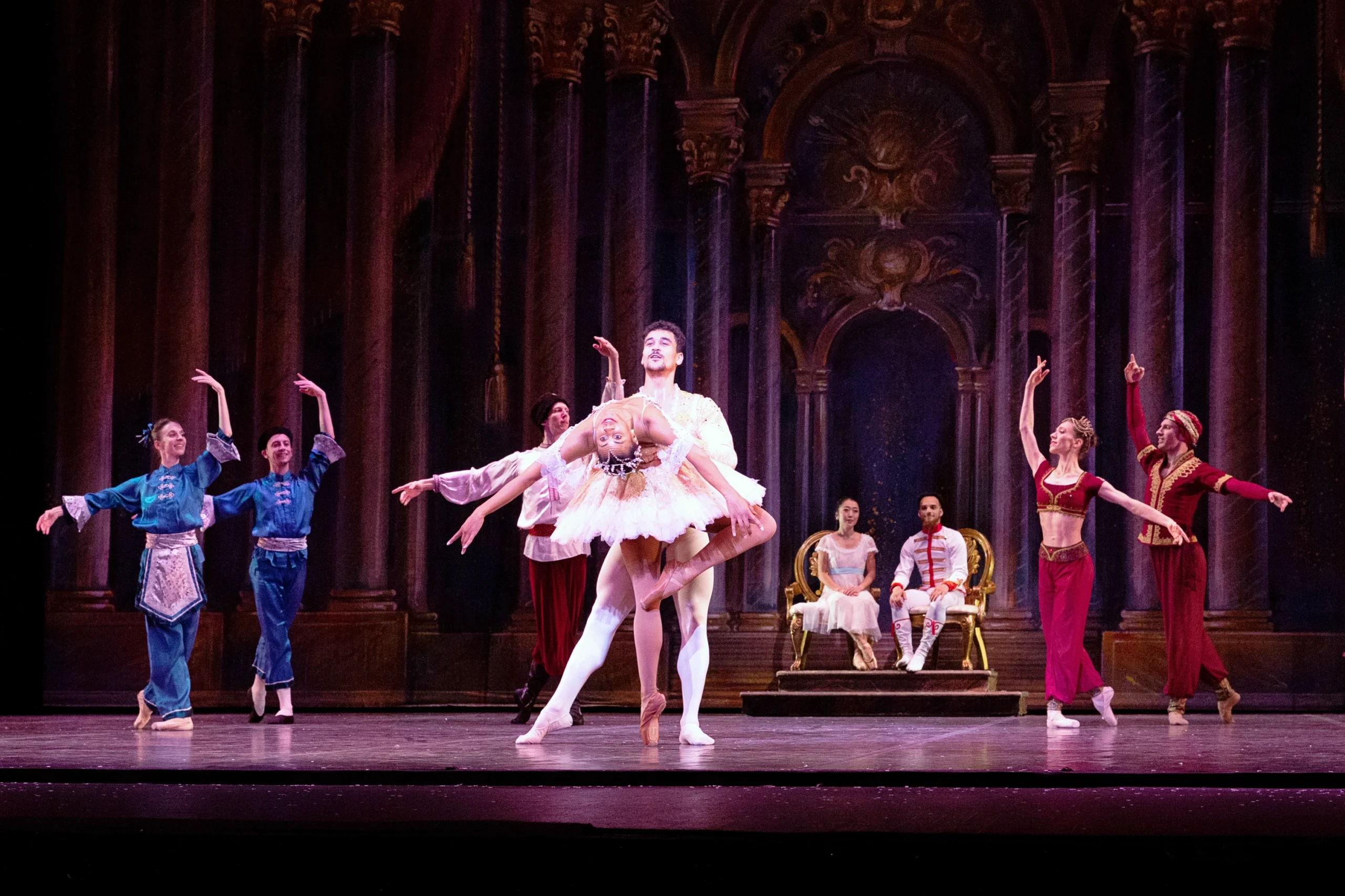 A large cast of dancers poses during a performance of Nutcracker. A ballerina portraying the Sugar Plum Fairy and her male partner stand center stage—he holds her waist as she stands facing him on pointe and does a deep backbend with her left leg in passé. She wears a pink tutu, brown tights and brown pointe shoes, while he wears white tights and slippers, and a white tunic. A male and female dancer sit in gilded chairs upstage center, while other dancers in various costumes frame the Sugar Plum Fairy and her cavalier in a V shape formation, posed in B plus.