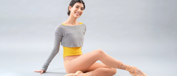 Jasmine Jimison sits on the floor against a light gray backdrop. She smiles towards the camera and crosses her right leg over her left, which is tucked underneath her, and points her right foot. She wears a yellow leotard under a gray, long-sleeved shrug, tan tights and tan pointe shoes.