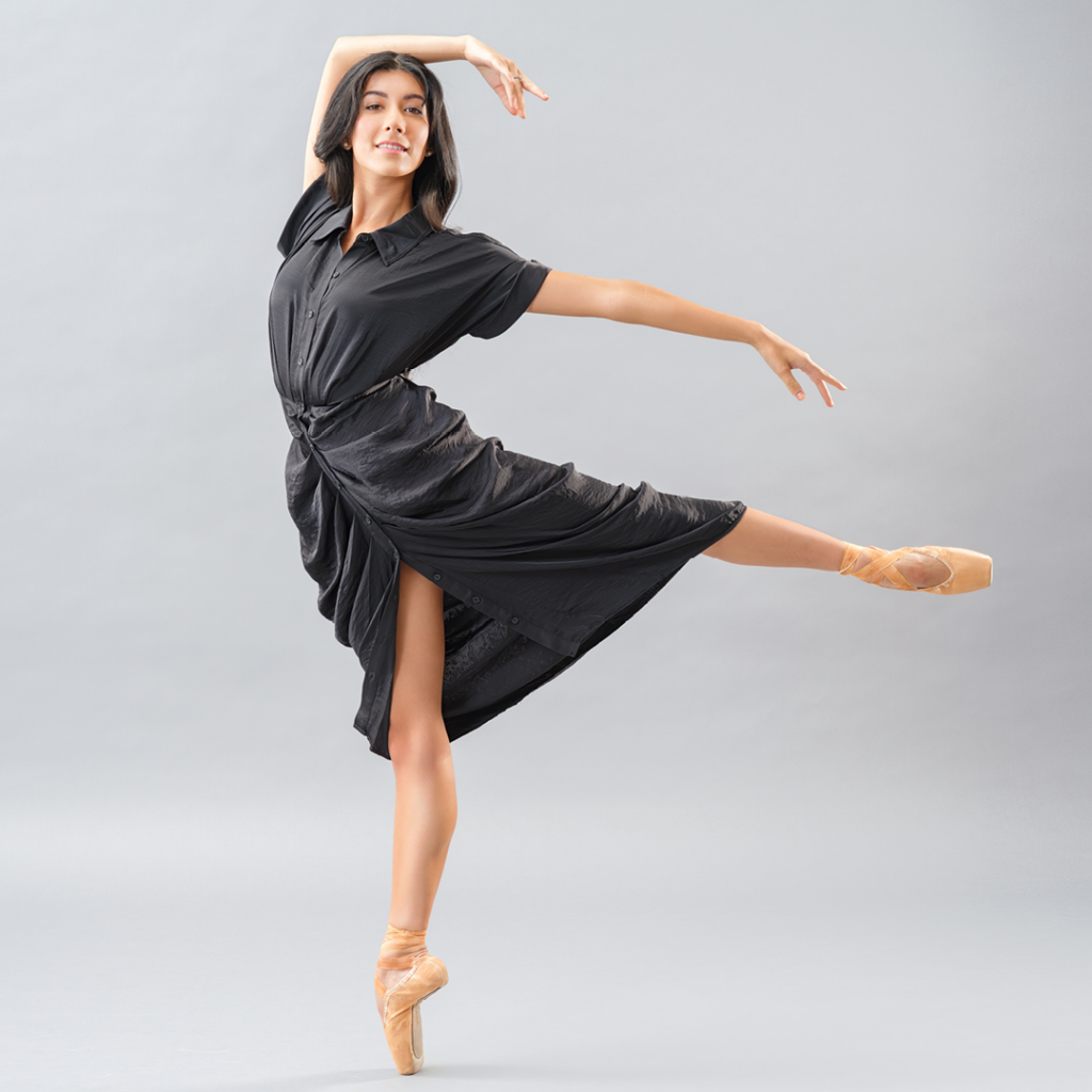 Jasmine Jimison poses in a low arabesque on pointe with her left leg lifted. She lifts her right arm and drapes it over her head and extends her left arm out to the side, and looks towards the camera wit a smile. She wears a shirtsleeved black, ruched dress and tan pointe shoes.