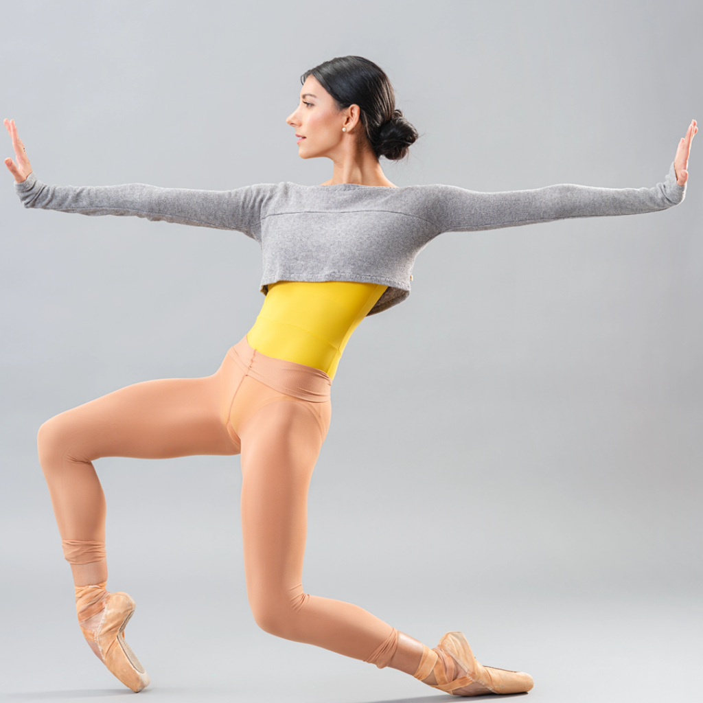 Jasmine Jimison is shown in profile in a wide stance, bending her knees and pushing over her pointe shoes. She reaches her arms out directly in front and back of her. She wears tan tights and pointe shoes, a yellow leotard and a long-sleeved gray sweater cropped at the ribs.