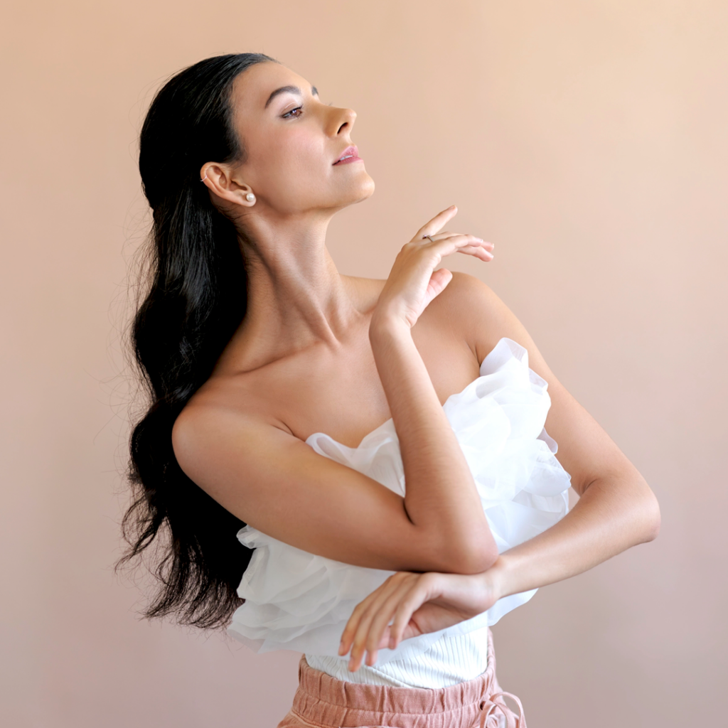Jasmine Jimison is shown in profile from the waist up wearing a strapless, fily white top and pink pants. She brings her right arm up, her hand just under her chin, and rests her left arm below it. Her dark hair is half pulled back at the top and falls down her back.
