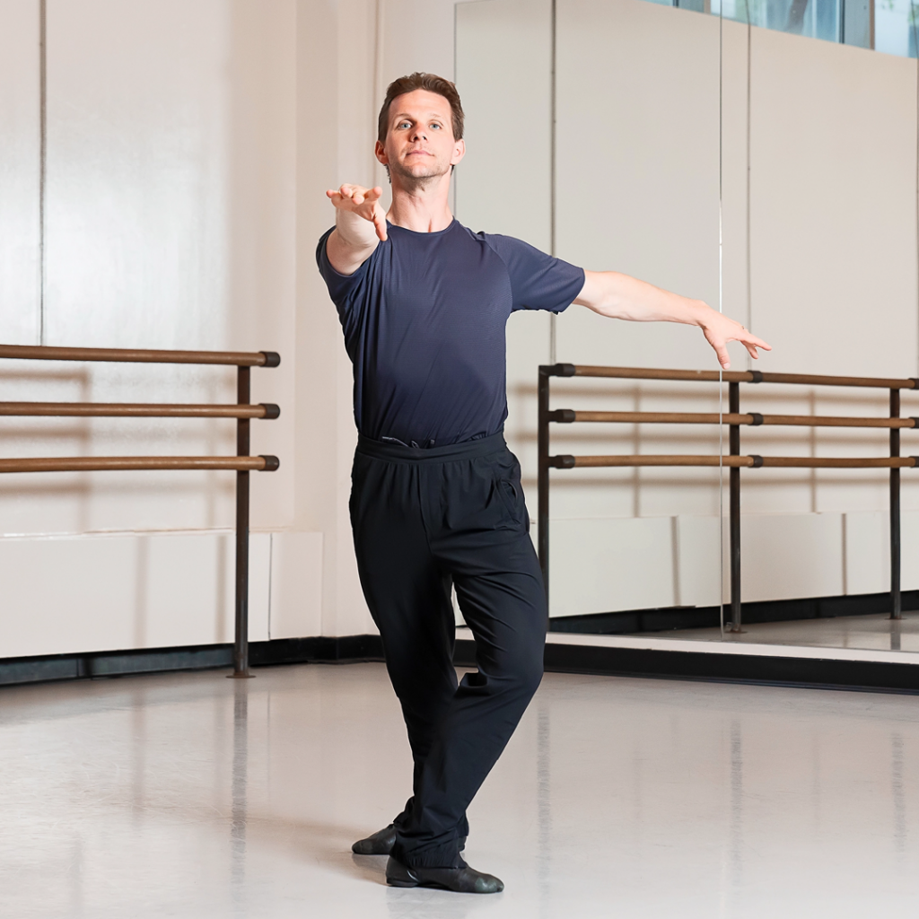 Daniel Ulbricht demonstrates a preparation for an à la seconde turn from fourth position with a straight back leg. He is in a ballet studio and wearing a black T-shirt with black pants and shoes.