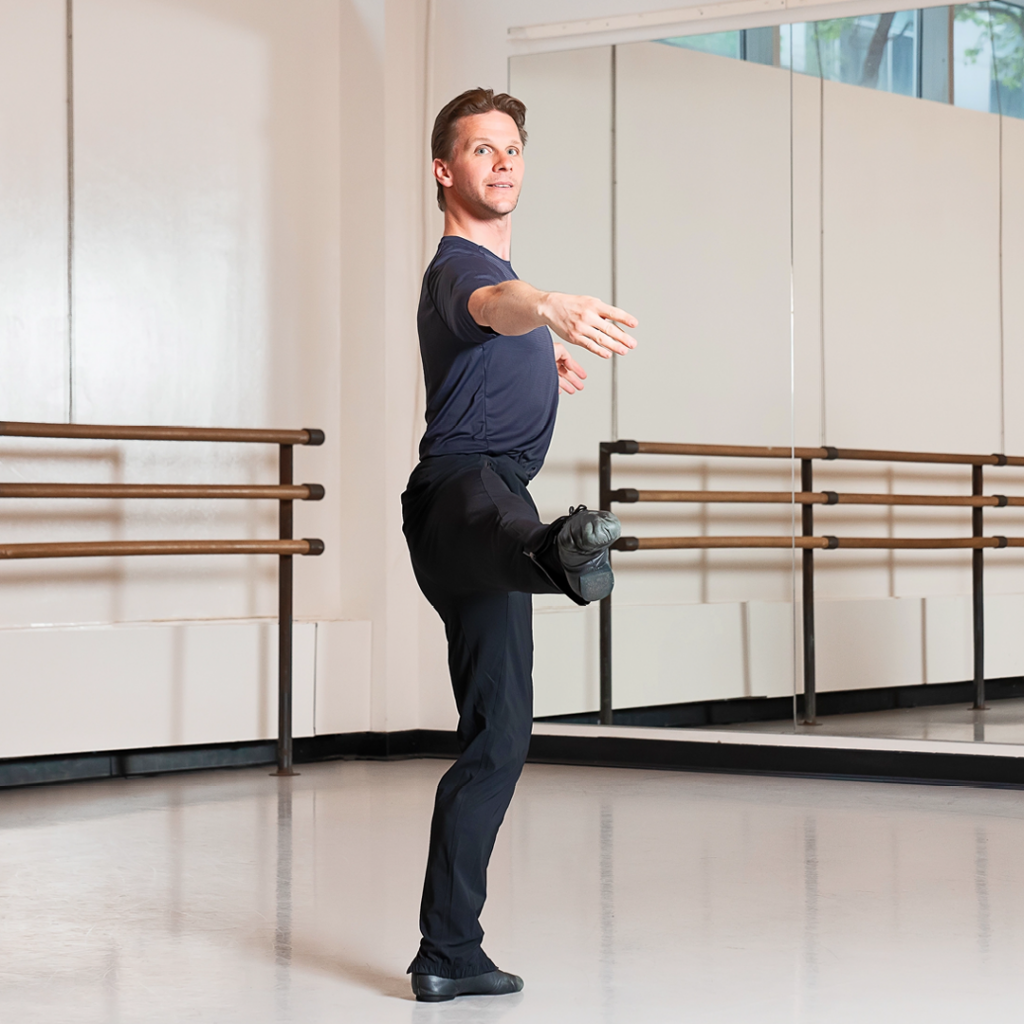 A side view of Daniel Ulbricht demonstrating plié between à la seconde turns, showing proper alignment. He is in a ballet studio and wearing a black T-shirt with black pants and shoes.