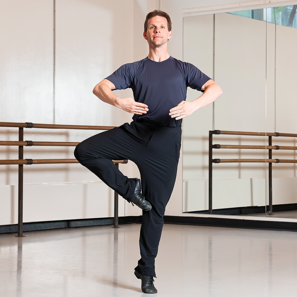 Daniel Ulbricht is demonstrating the pull-in portion of an à la seconde turn sequence. He is in a ballet studio and wearing a black T-shirt with black pants and shoes.