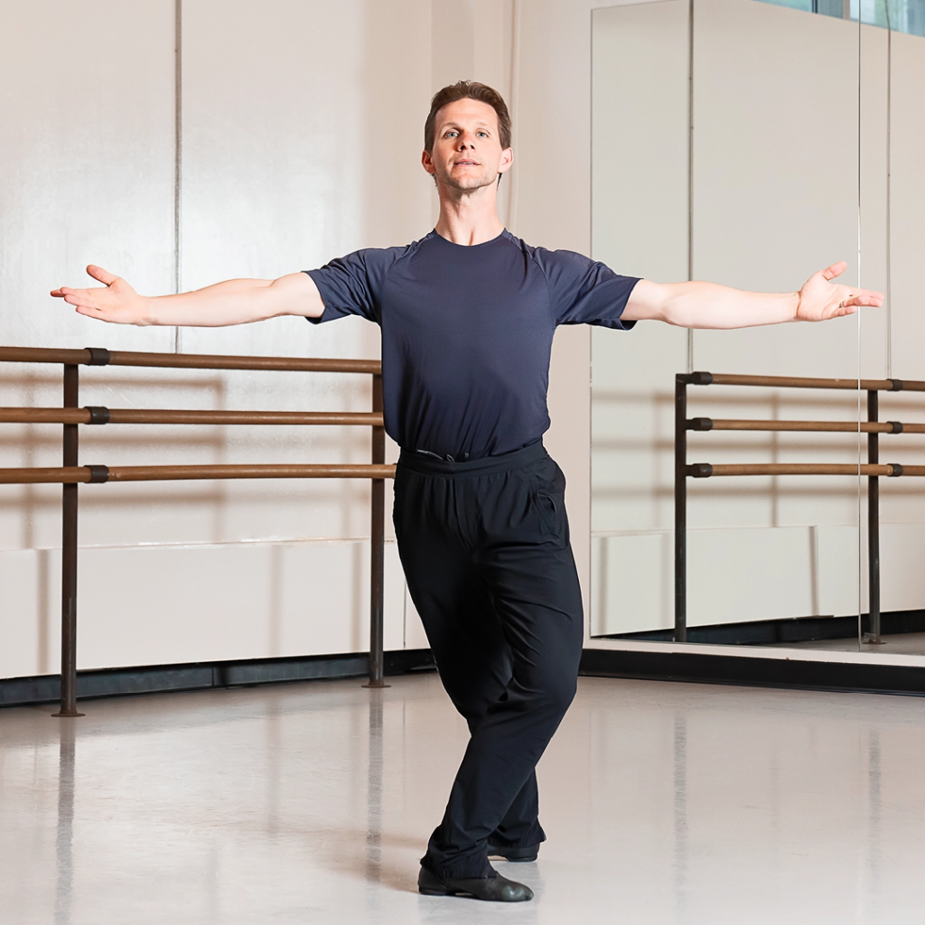 Daniel Ulbricht is demonstrating the finish after à la seconde turns. He is in a ballet studio and wearing a black T-shirt with black pants and shoes.