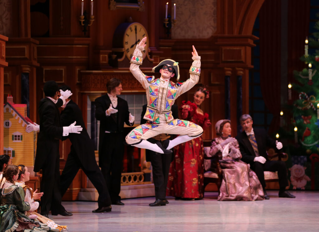 Paul Piner wears a Harlequin costume and jumps straight up with his legs turned out and tucked up underneath him during a performance of the Nutcracker. He wears a multi-colored Harlequin costume with white tights and black ballet slippers and holds his arms up, his elbows bent at right angles. Dancers in various party costumes mill about in the background and watch.