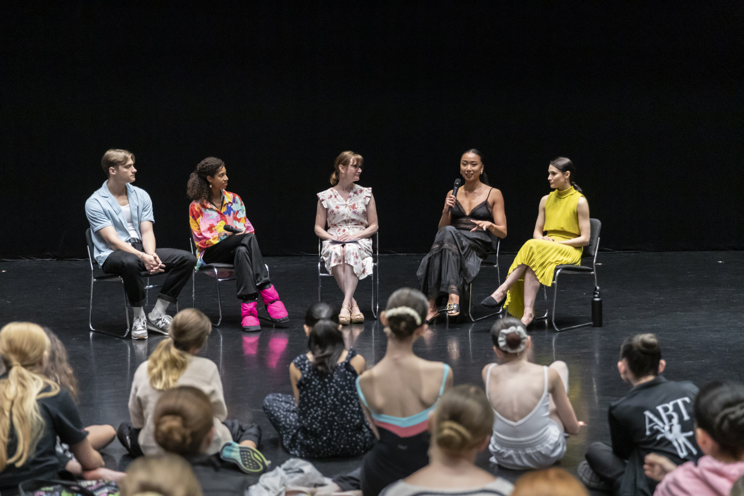 In a black box theater space, one man and four women sit in black chairs facing an audience of young ballet students sitting on the floor.