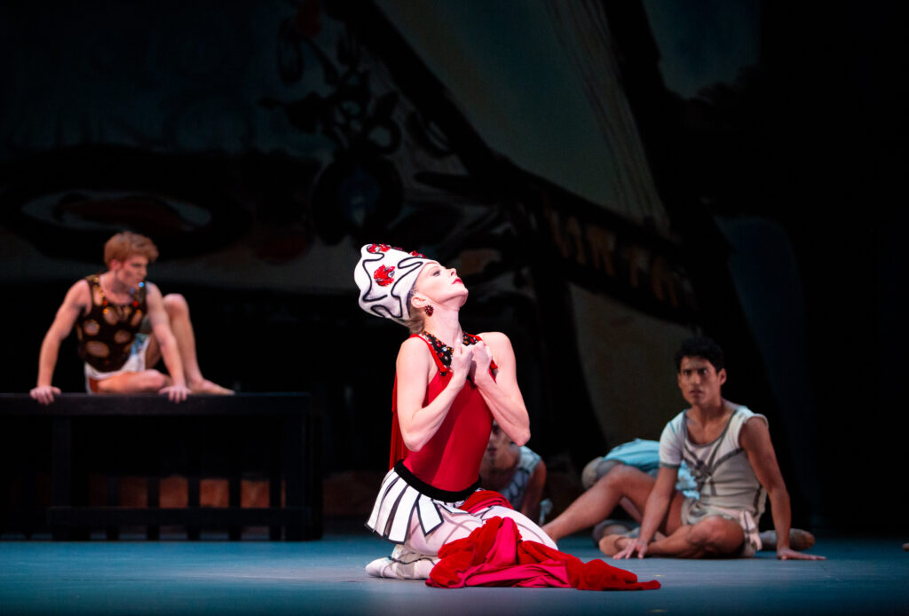 Dawn Atkins kneels onstage during a performance, looks up dramatically, and pulls her fists into her chest. She wears a tall white hat with red and black abstract designs, a short dress with a red bodice, pleated white skirt and long red cape, white tights and pointe shoes. The cape is draped around her left leg with the train piled in front of her. Behind her, two men sit onstage and watch her.