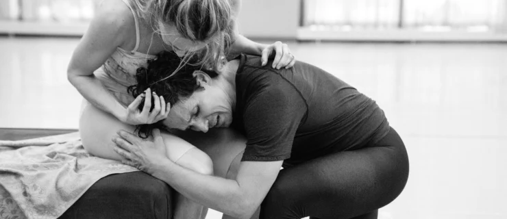 In this black and white photo, Sara Mearns and Guillaume Côté rehearse a scene from the ballet Romeo and Juliet in a large, brightly lit dance studio. Mearns, in a light colored leotard and cut-off pink tights, sits on a piano bench and huddles over Côté as he embraces her passionately and lays his head in her lap. Côté wears a dark T-shirt and black tights.