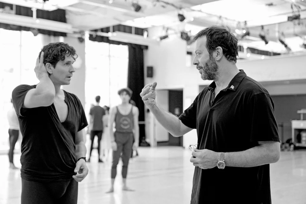 In this black and white photo, Alexei Ratmansky gives notes to Guillaume Côté in a large, brightly lit dance studio. They both wear dark shirts. Côté touches his hair with his right hand.