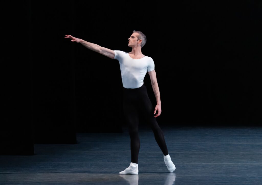 Russell Janzen stands onstage in a tendu in ecarte derriere, his right arm reaching out on a slight diagonal in front of him and his left resting gently at his side. He wears black ballet tights, a tight white cap-sleeve shirt, white socks, and white ballet shoes.