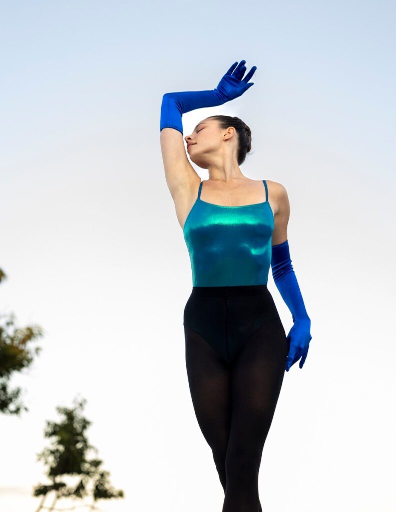 Sierra Armstrong, shown knees-up from below, performs in a bright, shiny teal leotard and black tights, with long cobalt blue gloves up past her elbows. She dances outdoors at Hamptons Dance Festival in front of a blue sky and lifts her right arm up, nestling her face gently.