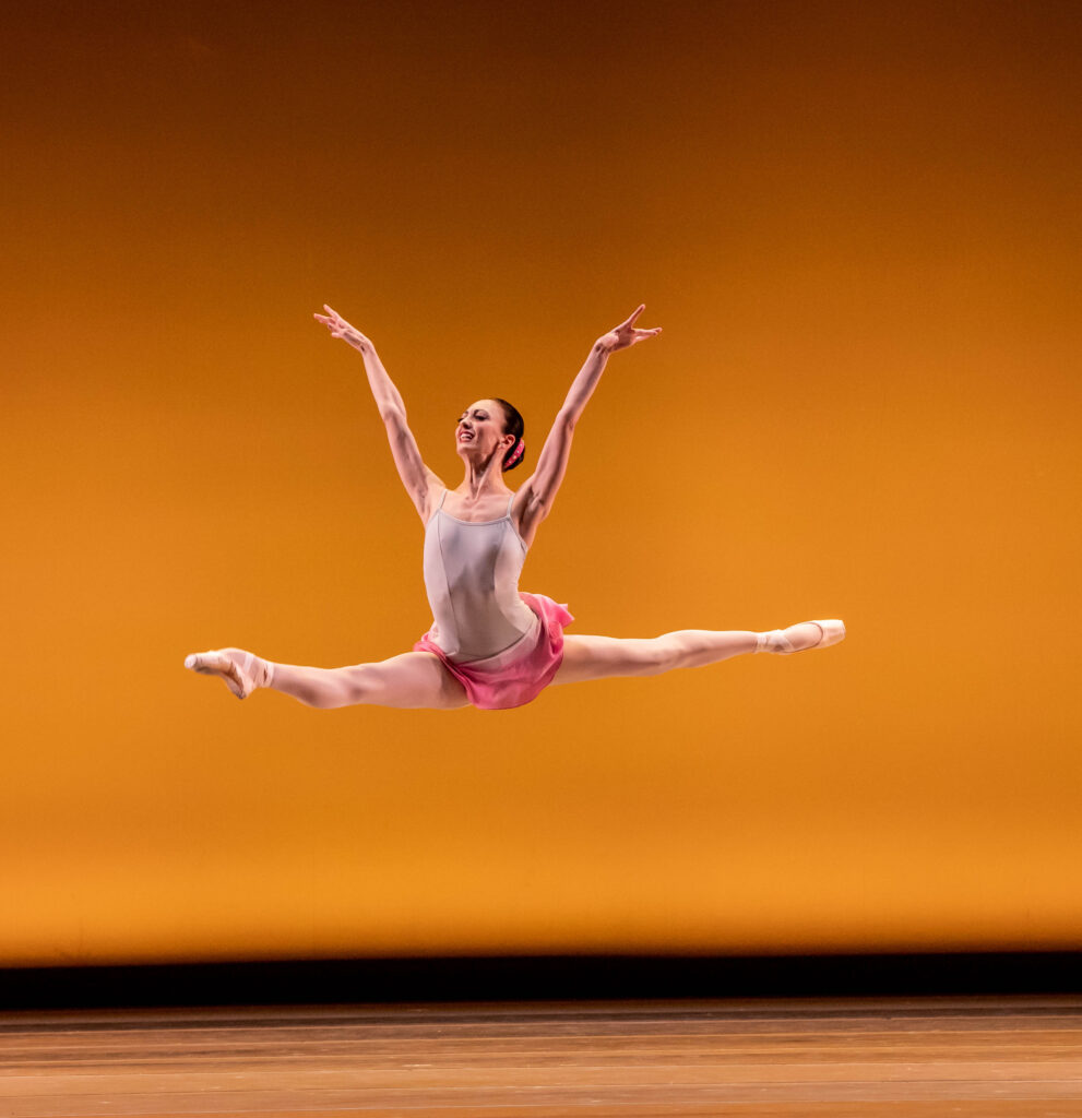 a ballerina flies mid–saut de chat onstage in front of an orange backdrop, lifting her arms in a "V" shape.