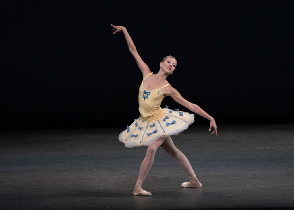 Dawn Atkins takes a wide fourth position lunge onstage with her left leg in front. She reaches her right arm high and her left arm low and looks out towards the audience, smiling. She wears a yellow tutu with blue bows on the skirt and bodice, pink tights and pointe shoes, and dances in front of a black backdrop.