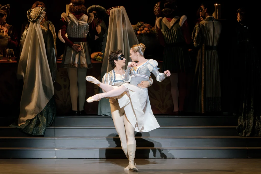 During a performance of the ballet Romeo and Juliet, Guillaume Côté carries Sara Mearns in his arms, her legs outstretched, as a large cast of dancers behind them face upstage, milling about and talking around a large table of food. The pair look at each other adoringly. Côté wears an off-white tunic, tights and ballet boots, his eyes covered by a black mask. Mearns wears a gray and white empire-waisted dress, pink tights and pointe shoes. Her hair is styled in an elaborate bun with ribbons and braids.