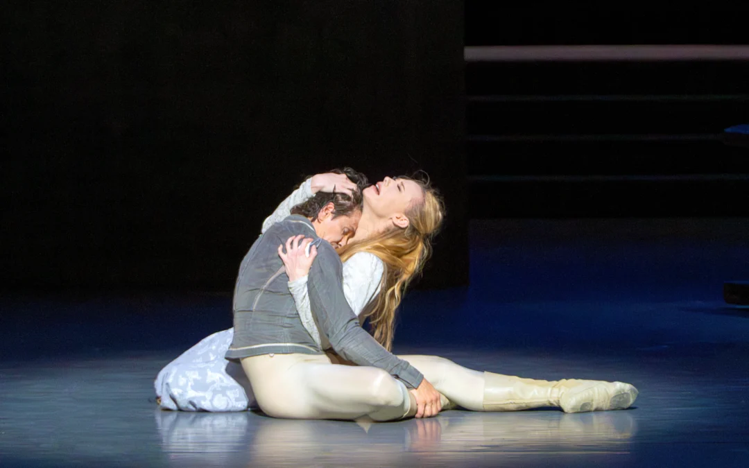 During a ballet performance of Romeo and Juliet, Sara Mearns sits on the stage facing stage right, cradling a limp Guillaume Côté in her arms. She leans back with her eyes closed, her face in anguish. She wears a light blue patterned dress, while Côté is costumed in a gray tunic and off-white tights and ballet boots.
