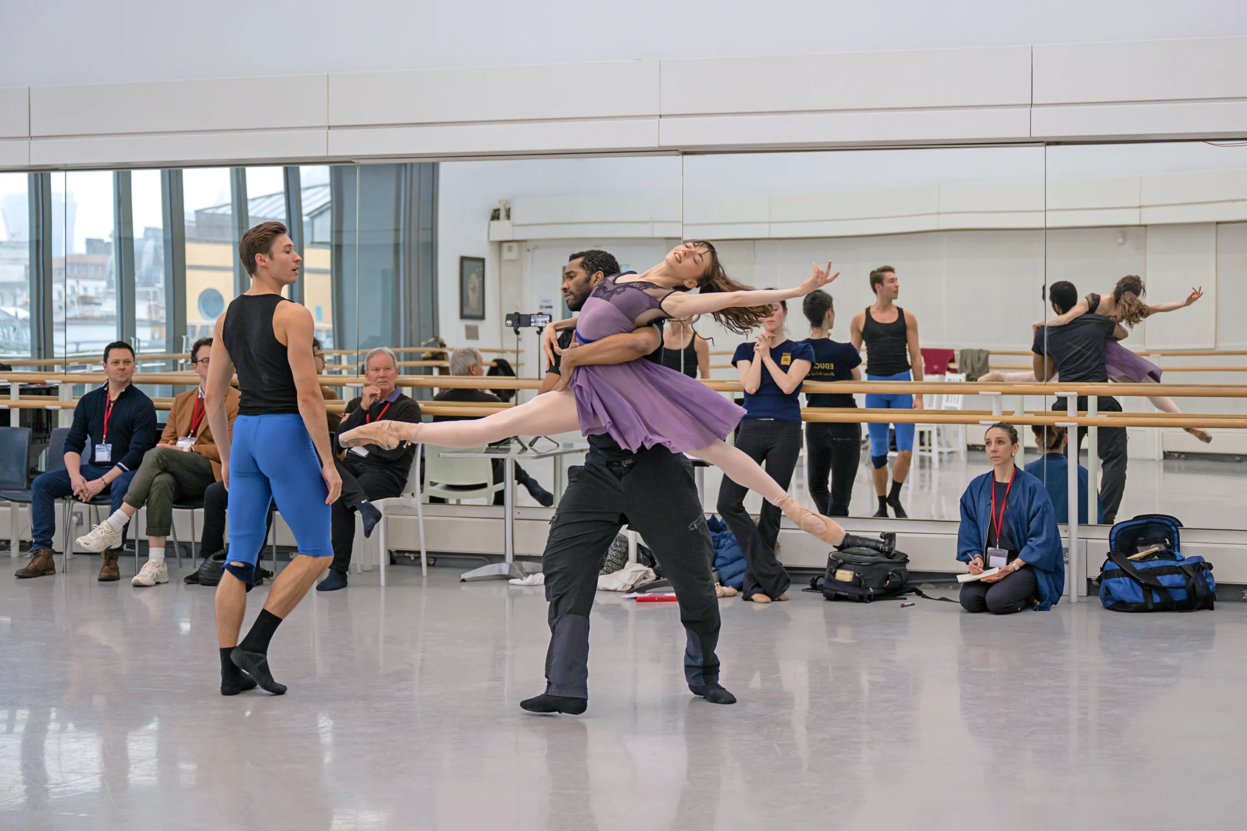 In a large dance studio, a group of dancers and rehearsal directors watch three dancers rehearse. A male dancer lifts a female dancer in a spinning lift, her legs in a split, and she arches backward. A second male dancer watches and walks close by.