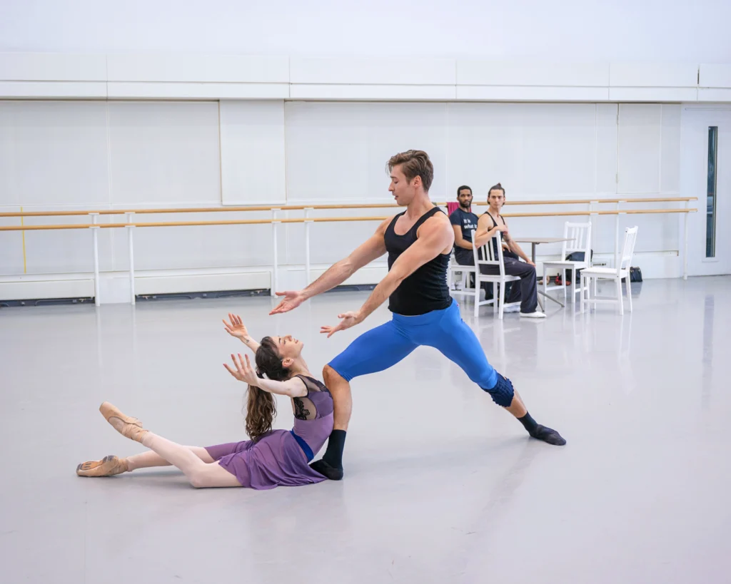 Anabelle de la Nuez and Graham Maverick rehearse together, with two male dancers sitting in chairs and watching in the background. Anabelle arches back in a low attitude on the floor, facing Graham, who lunges forward and reaches toward her