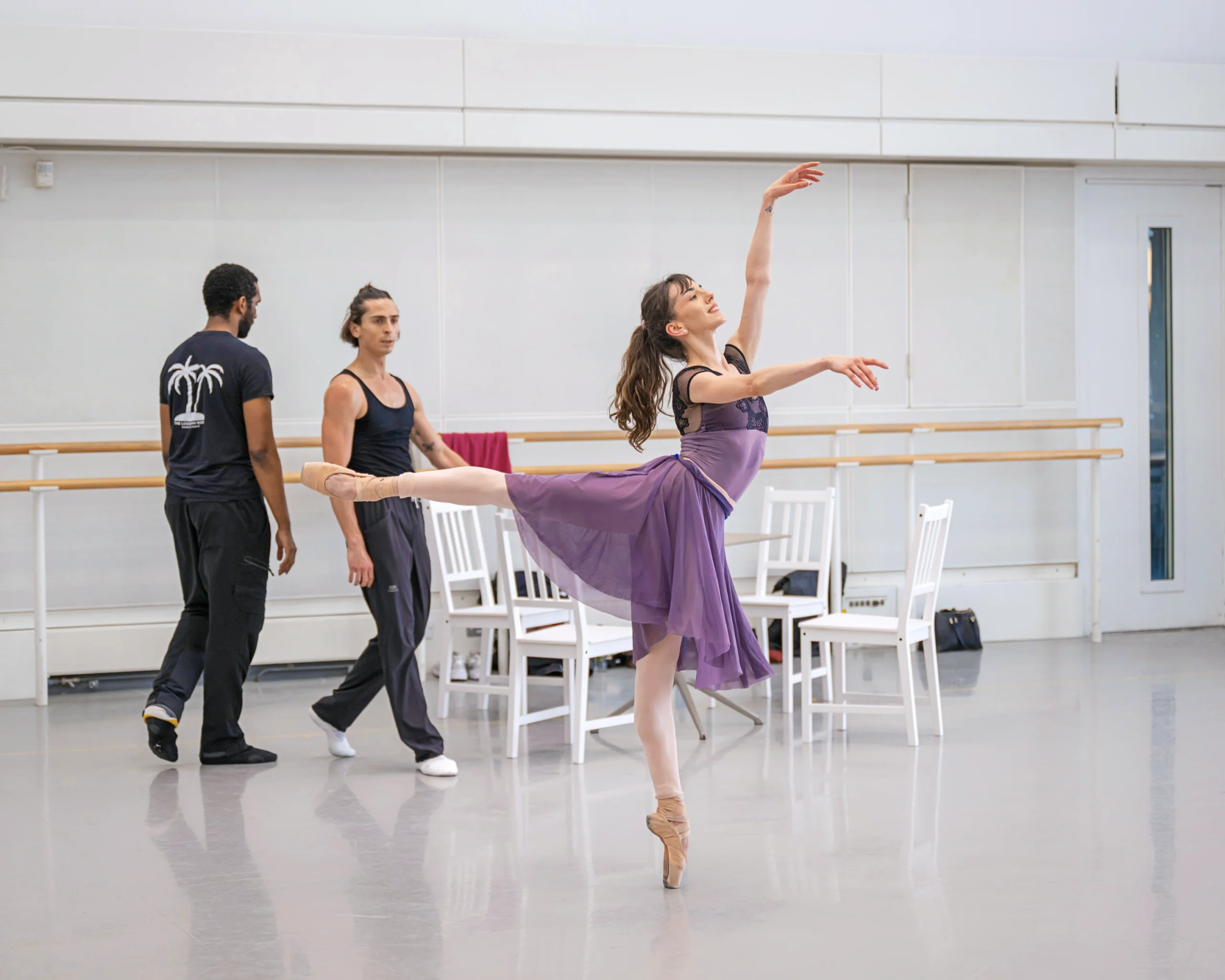 In a large dance studio, a female dancer does an arabesque on pointe in front of two male dancers, who walk behind her next to four white chairs.