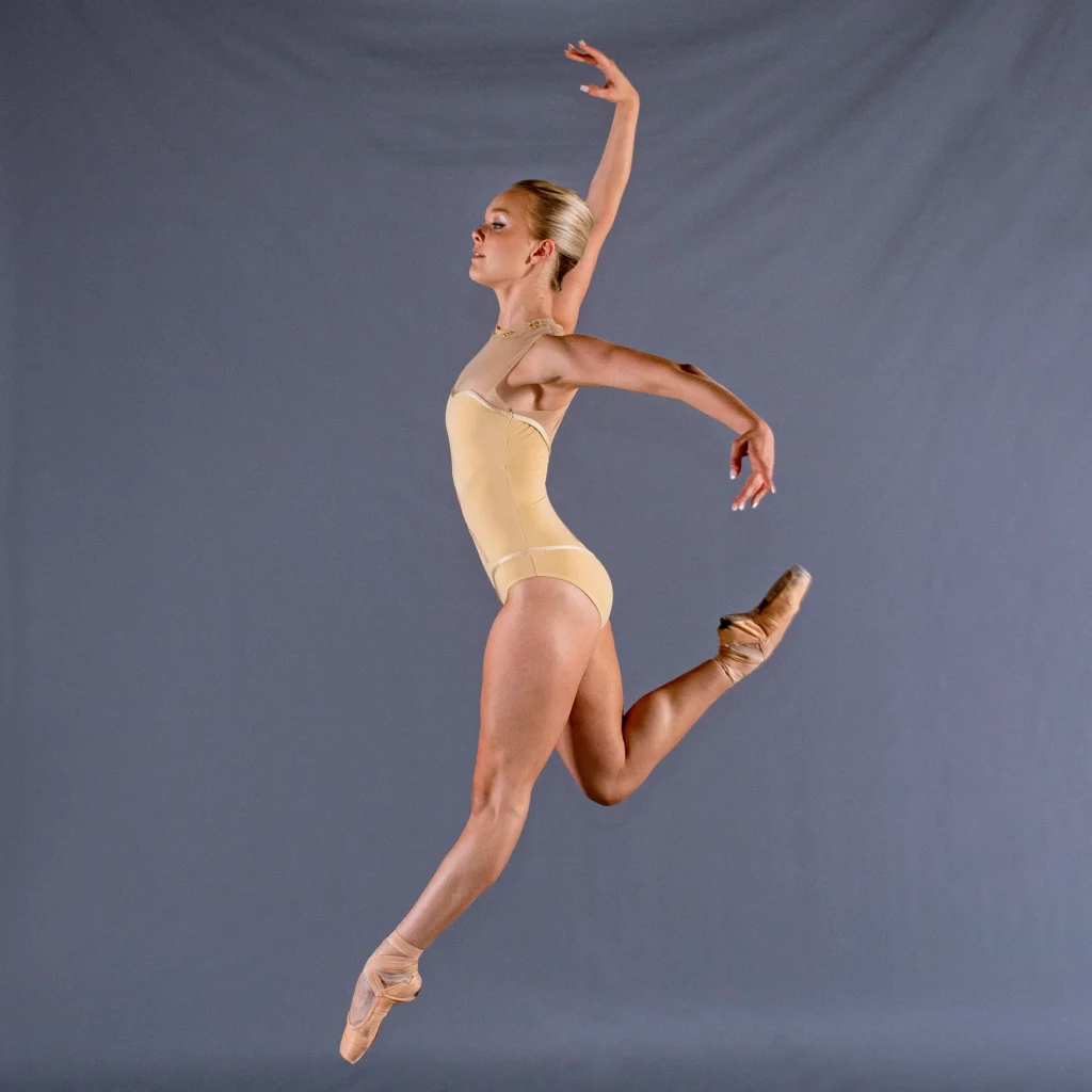 Adriana Wagenveld, shown in profile, does a small jump in front of a gray backdrop. She tretches her left leg out and tucks her right leg up behind her, lifting her right arm out and her left arm to the side and slightly back. She wears a yellow leotard and tan pointe shoes, and smiles slightly.