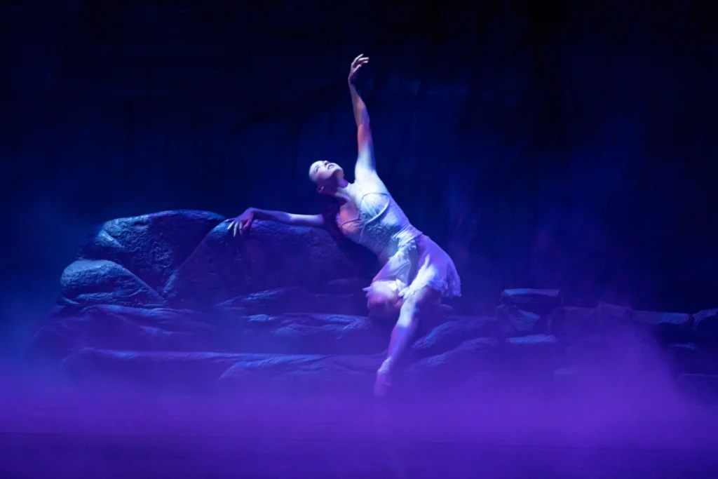 Onstage amid hazy purple-blue lighting, a female dancer sits on a large rock formation and reaches upward to the ceiling.