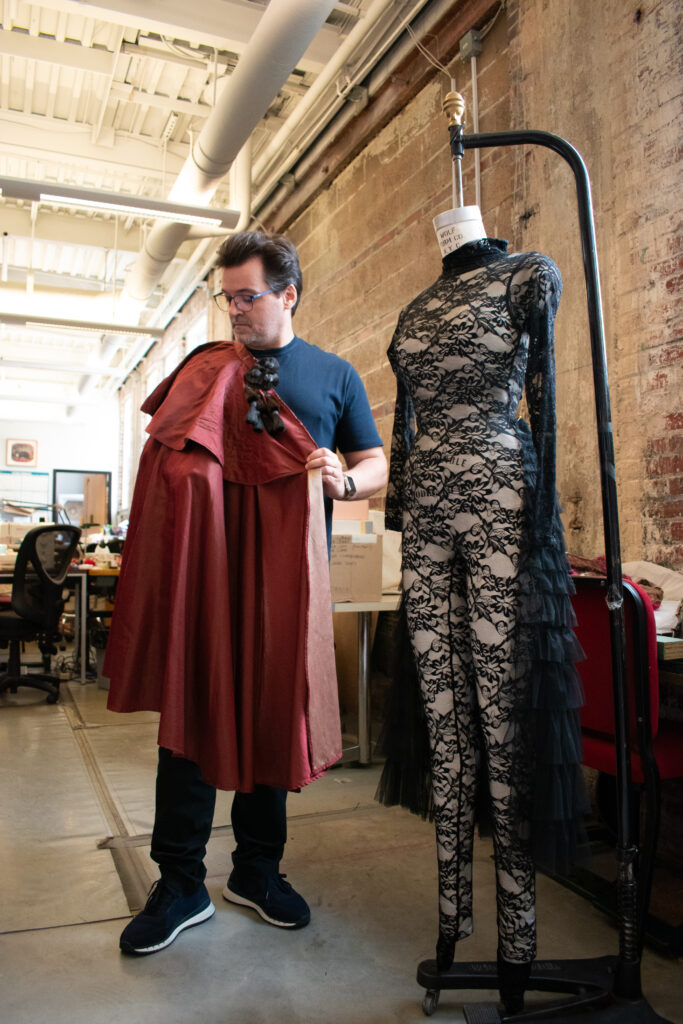 Angel Corella poses with costumes for his "Carmen" ballet, wearing a red toreador cape. Next to him, a black, lacy unitard is displayed on a mannequin.