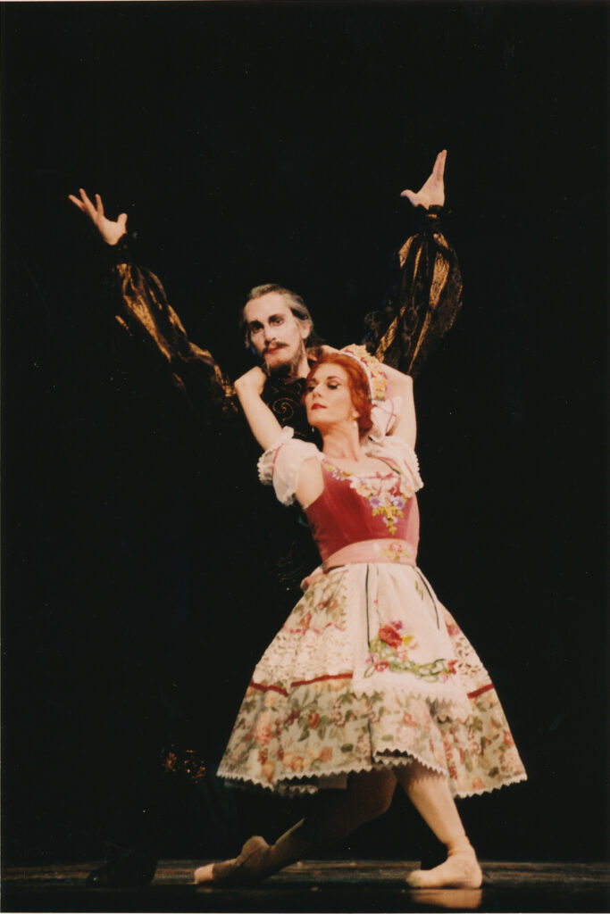 Tim O'Keefe performs as Dracula and dances a pas de deux with a female dancer playing Lucy. She hangs onto his neck from the front, looking up at him, and he stands with his arms in a V-shape.