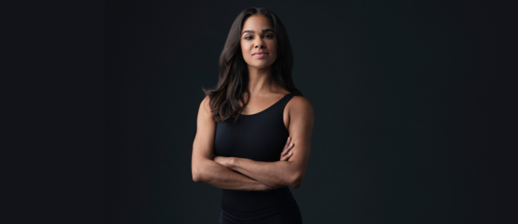 Misty Copeland stands with her arms crossed confidently and gives a sly smile as she poses for a portrait in front of a dark gray backdrop.