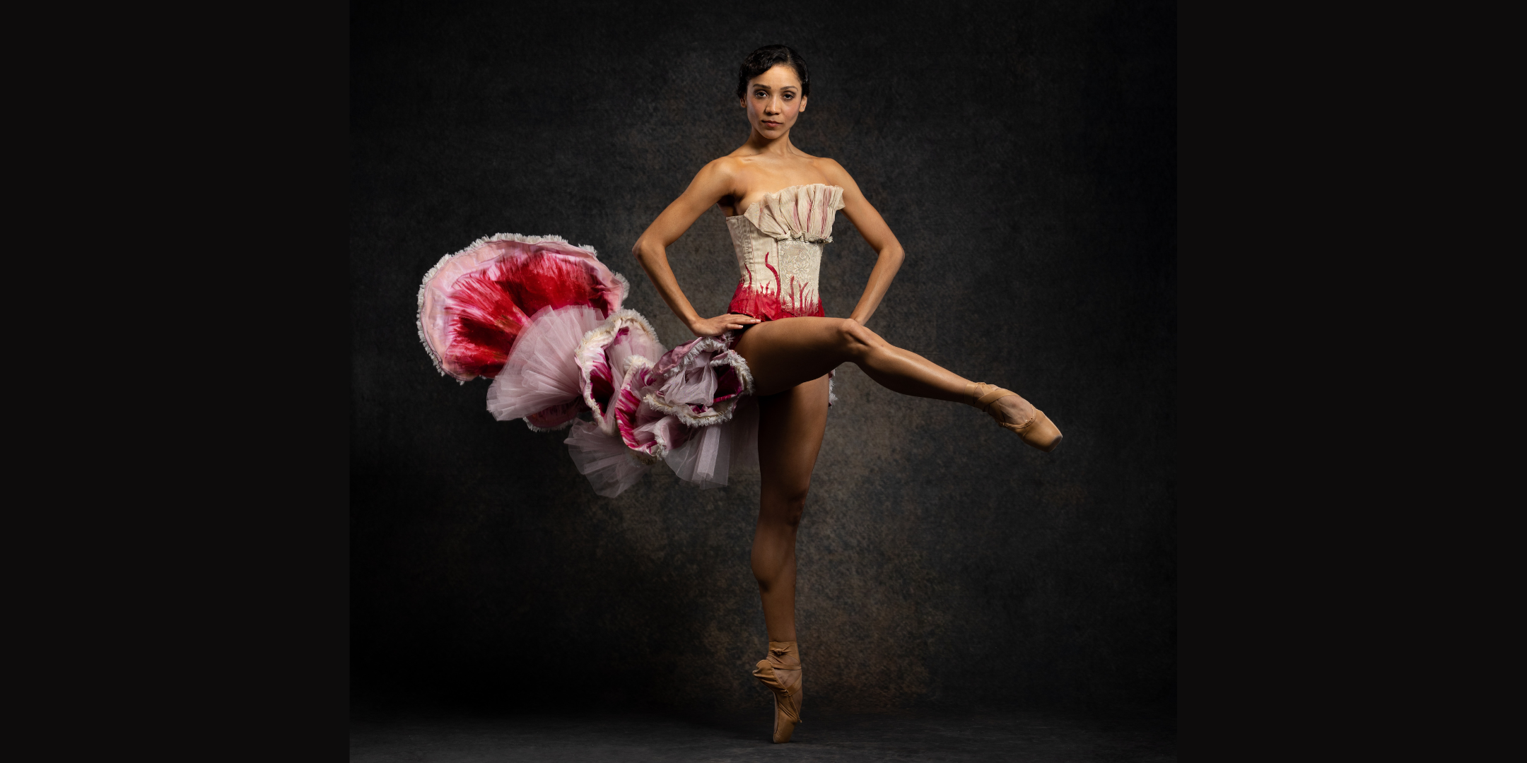 Nayara Lopes poses in a croisé attitude devant en pointe as Carmen. She wears a long, layered scarlet skirt with gold trimming and a gold bodice. She rests her hands on her hips and looks into the camera with a sly smile.